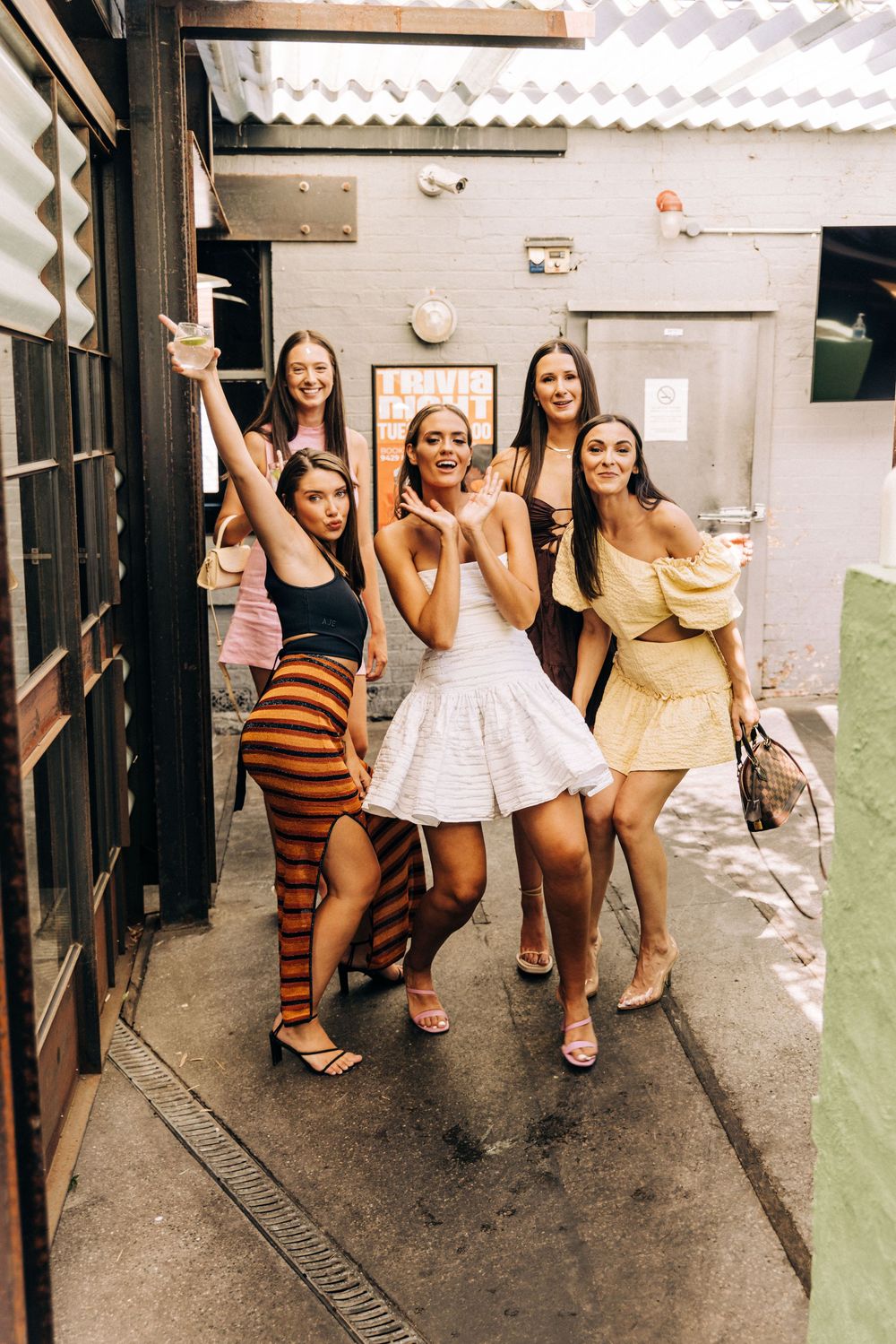 Five young women in trendy outfits posing playfully in an outdoor alleyway venue.