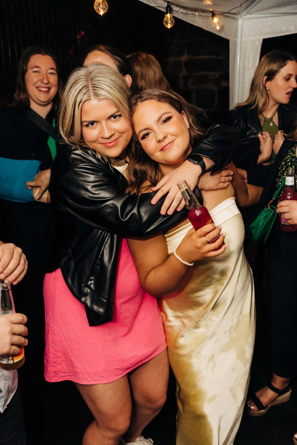 Two women hugging and smiling at a party, one in a pink dress with leather jacket, other in gold satin dress.