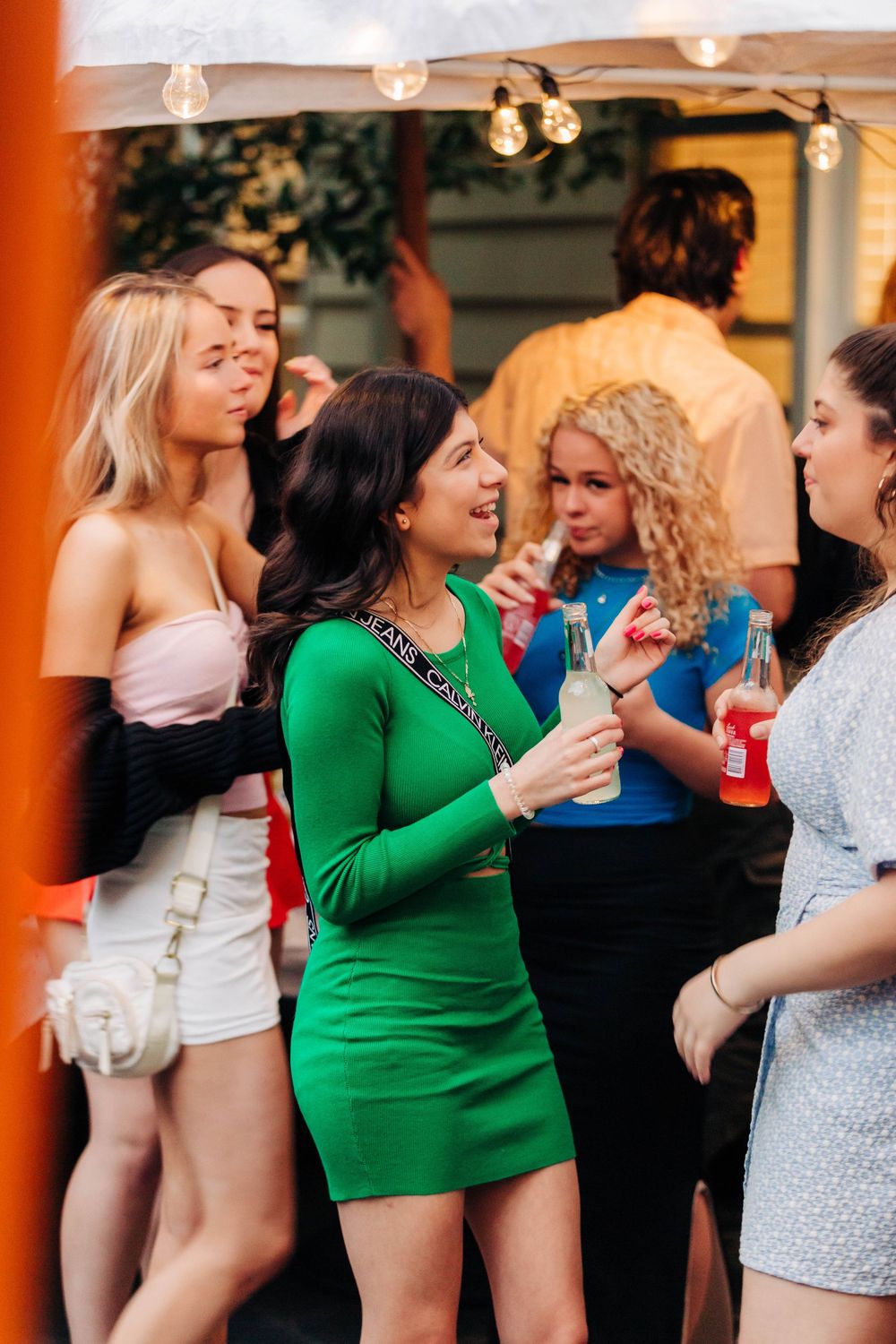 Group of young women socializing at an outdoor party, holding drinks, wearing colorful outfits under string lights.