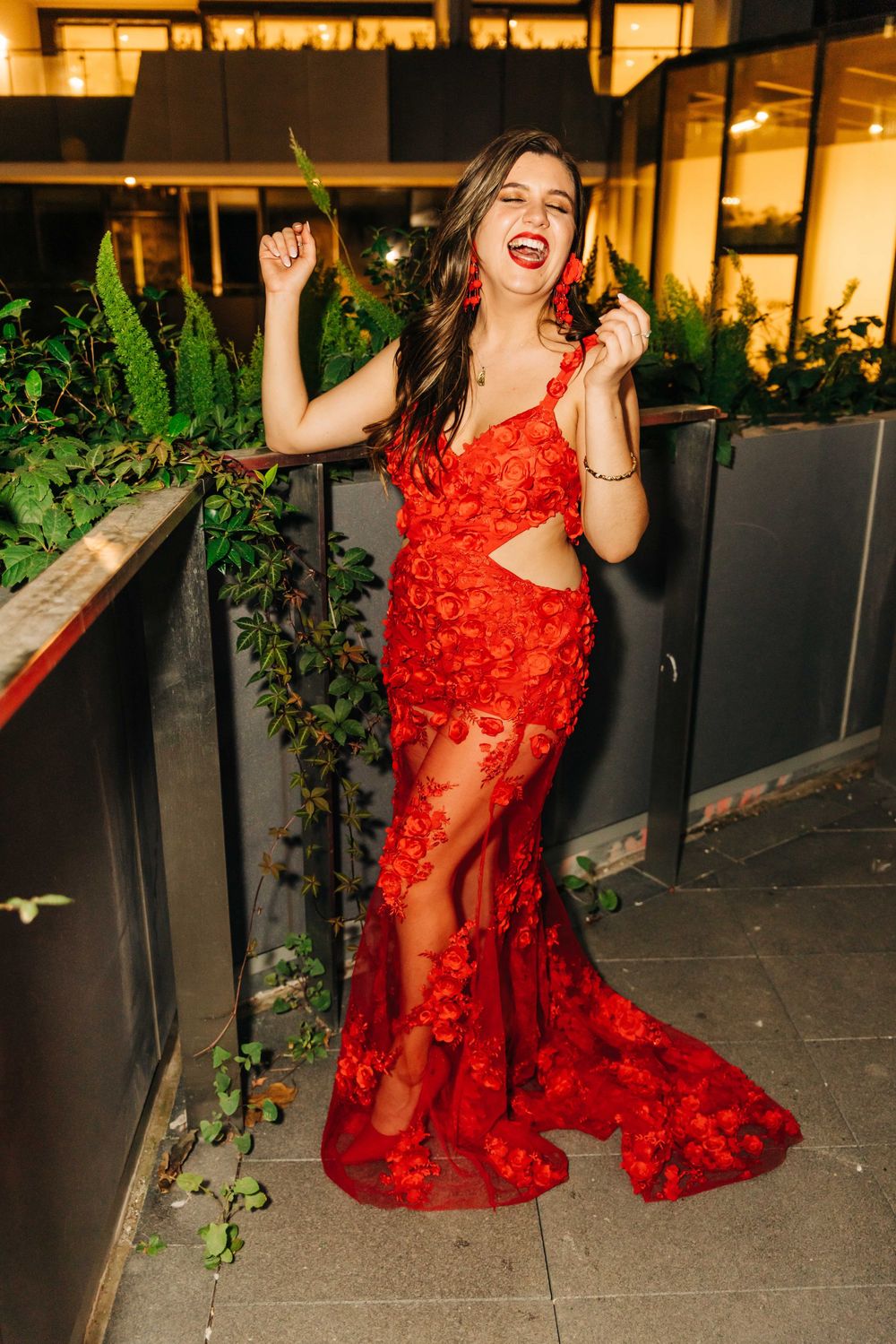 Woman in red floral lace gown with cutout waist laughing outdoors at night.