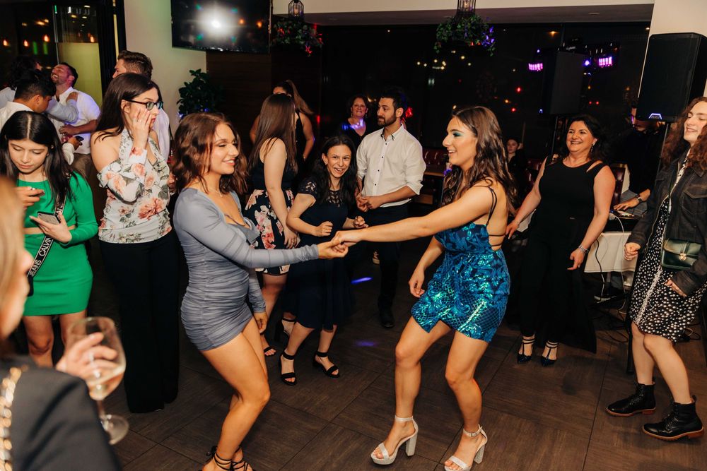 Two women dancing together at a lively party event with guests socializing in the background.