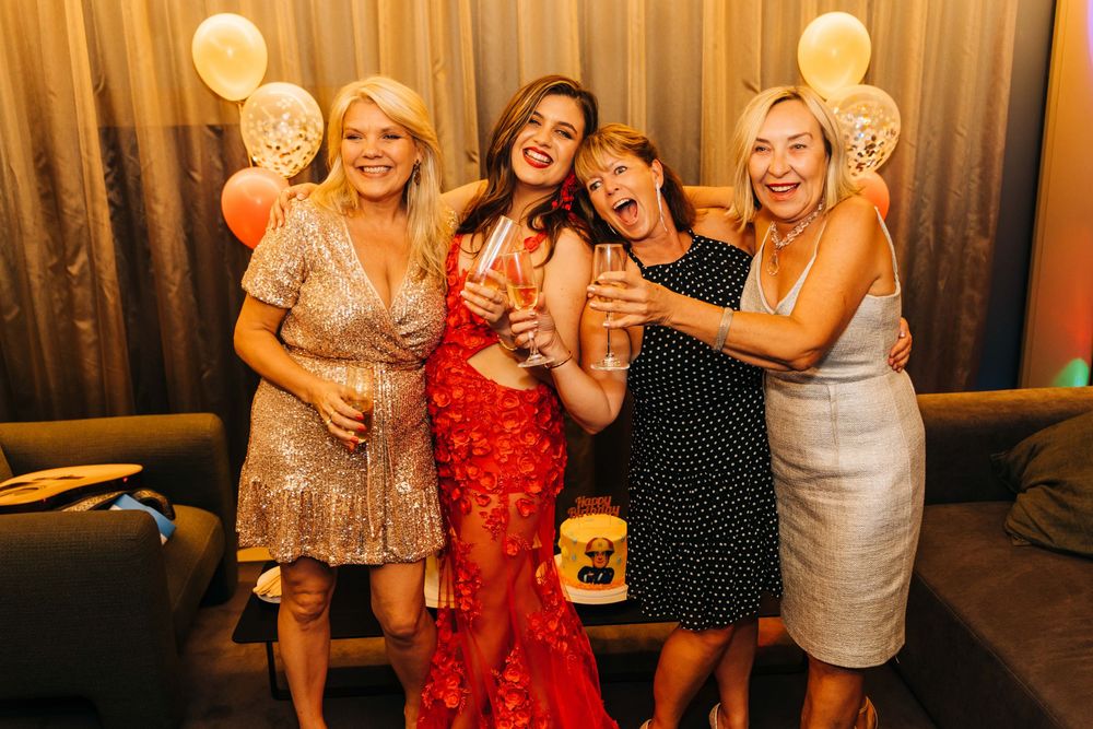 Four women smiling and celebrating at a party with balloons and champagne glasses in a festive venue.
