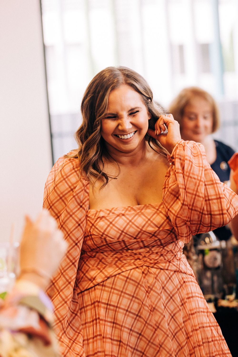 A smiling woman in an orange plaid dress touches her ear at an indoor event.