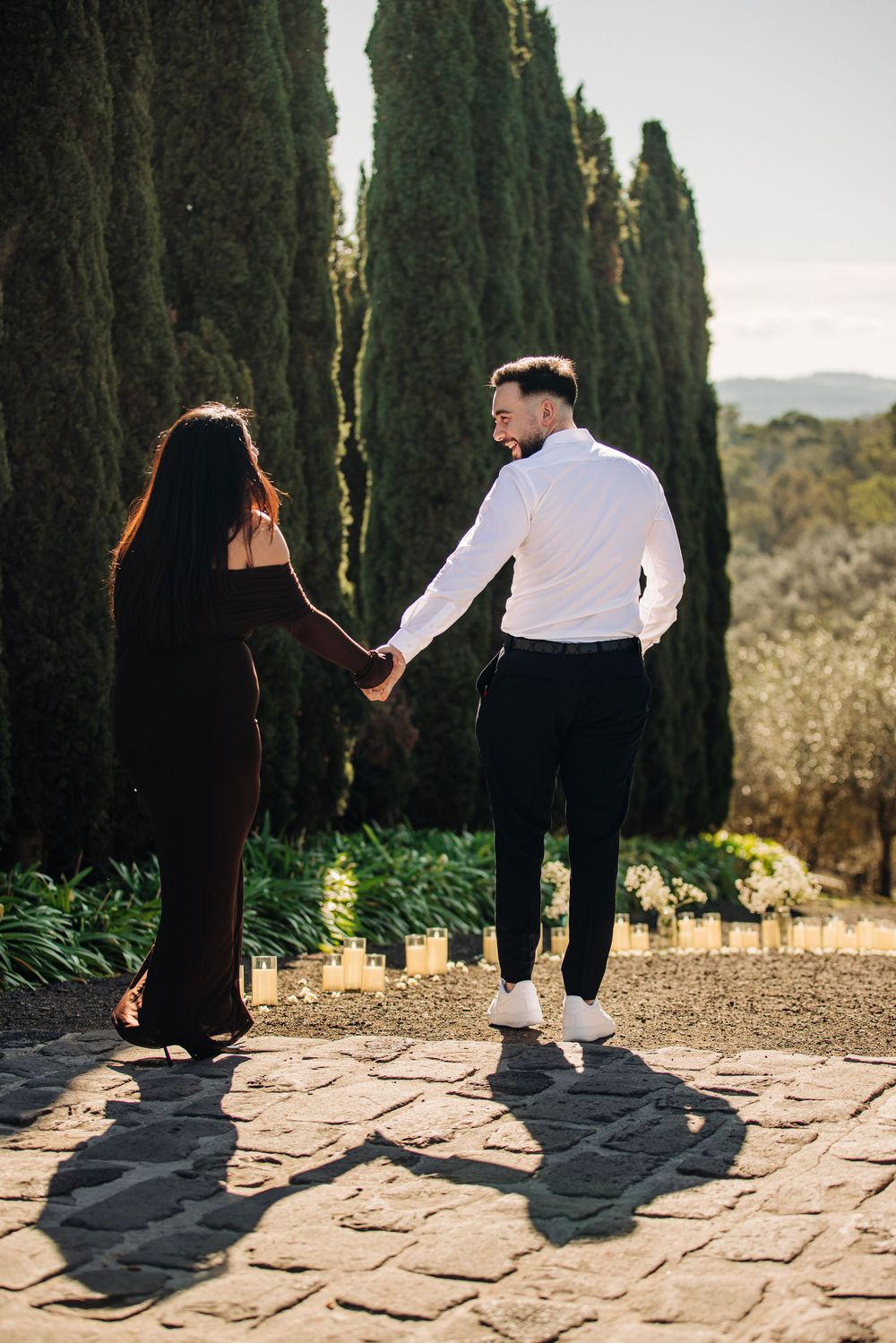 Couple holding hands on a stone path surrounded by tall cypress trees in a sunny outdoor setting.