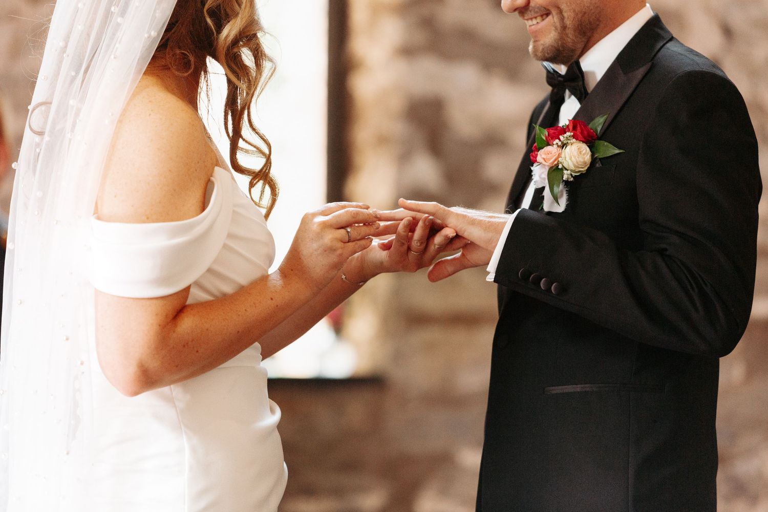 Bride placing a wedding ring on groom's finger during ceremony, groom wearing black tuxedo with floral boutonniere.
