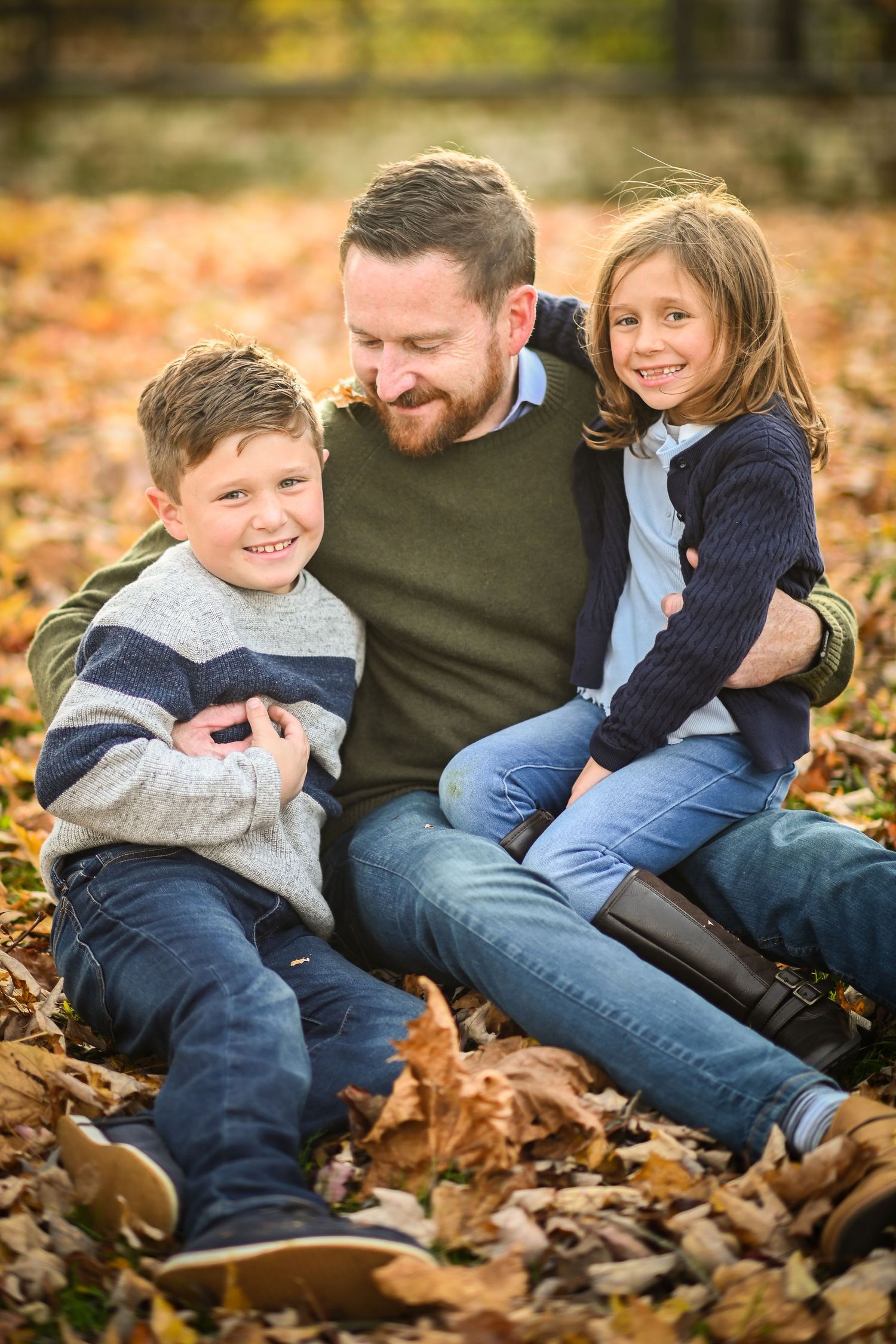 A bald father sits on grass with two daughters, laughing together amid colorful autumn foliage.