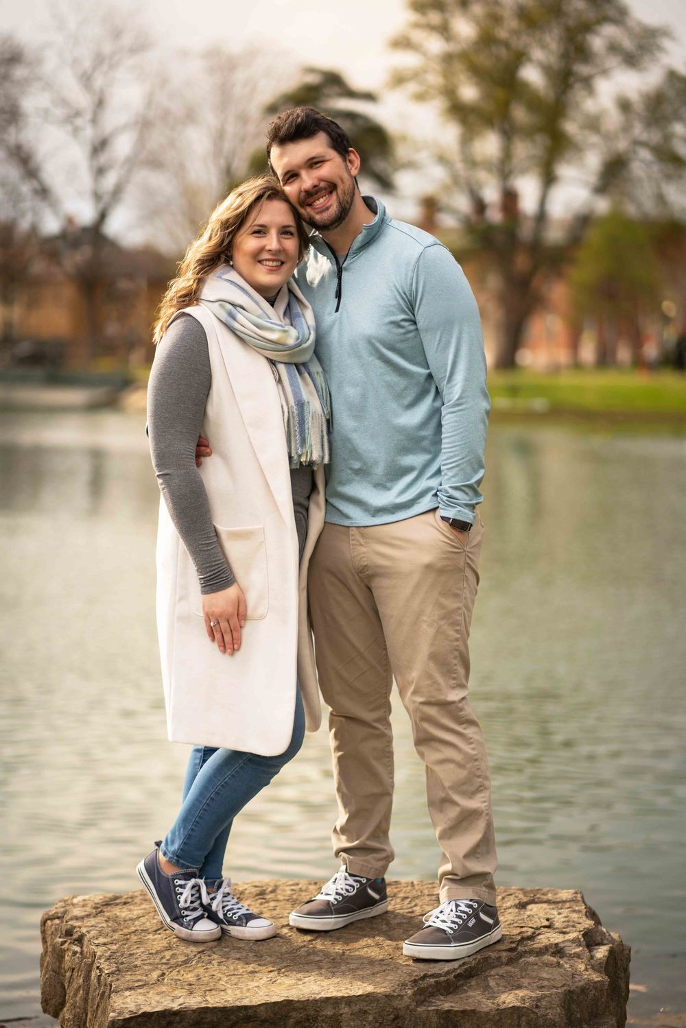 Smiling couple standing on a rock by a pond in a park, wearing casual fall outfits including scarves and sneakers.