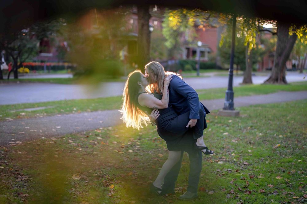 Couple sharing a romantic dip kiss in a lush green park, surrounded by trees and soft golden light.