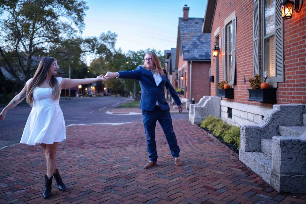 Couple dancing on brick pathway near historic building at dusk, woman in white dress and man in navy suit.