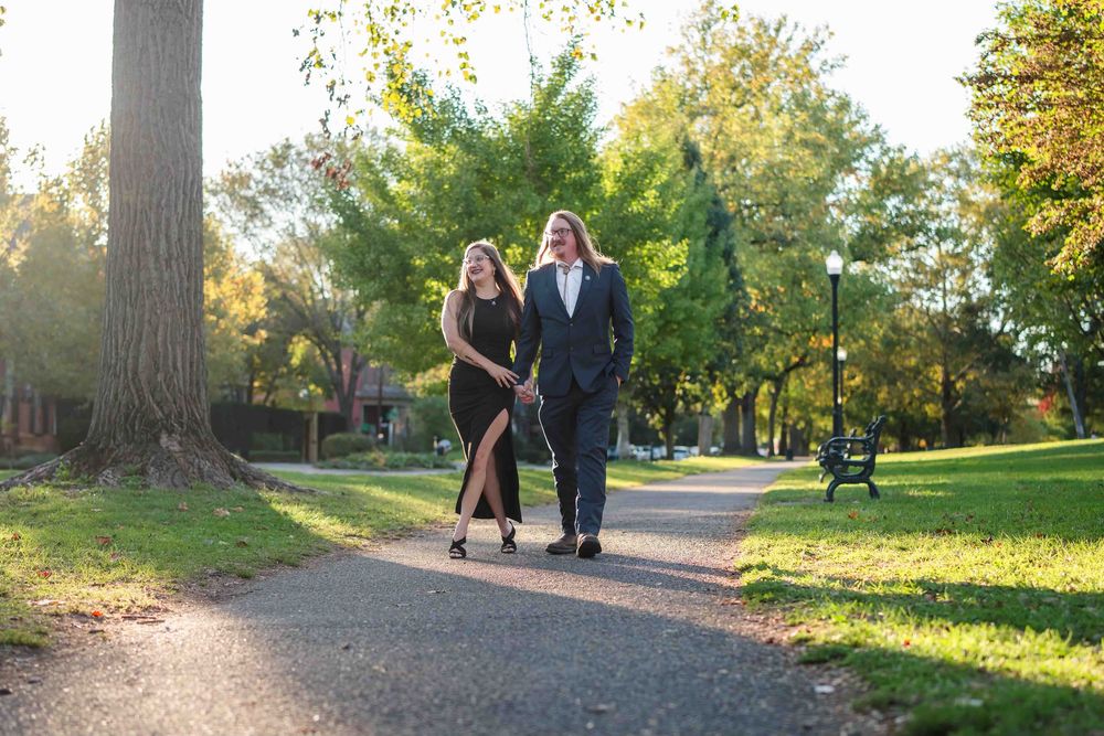 Couple in formal attire walking hand in hand on a sunlit park path surrounded by lush green trees.