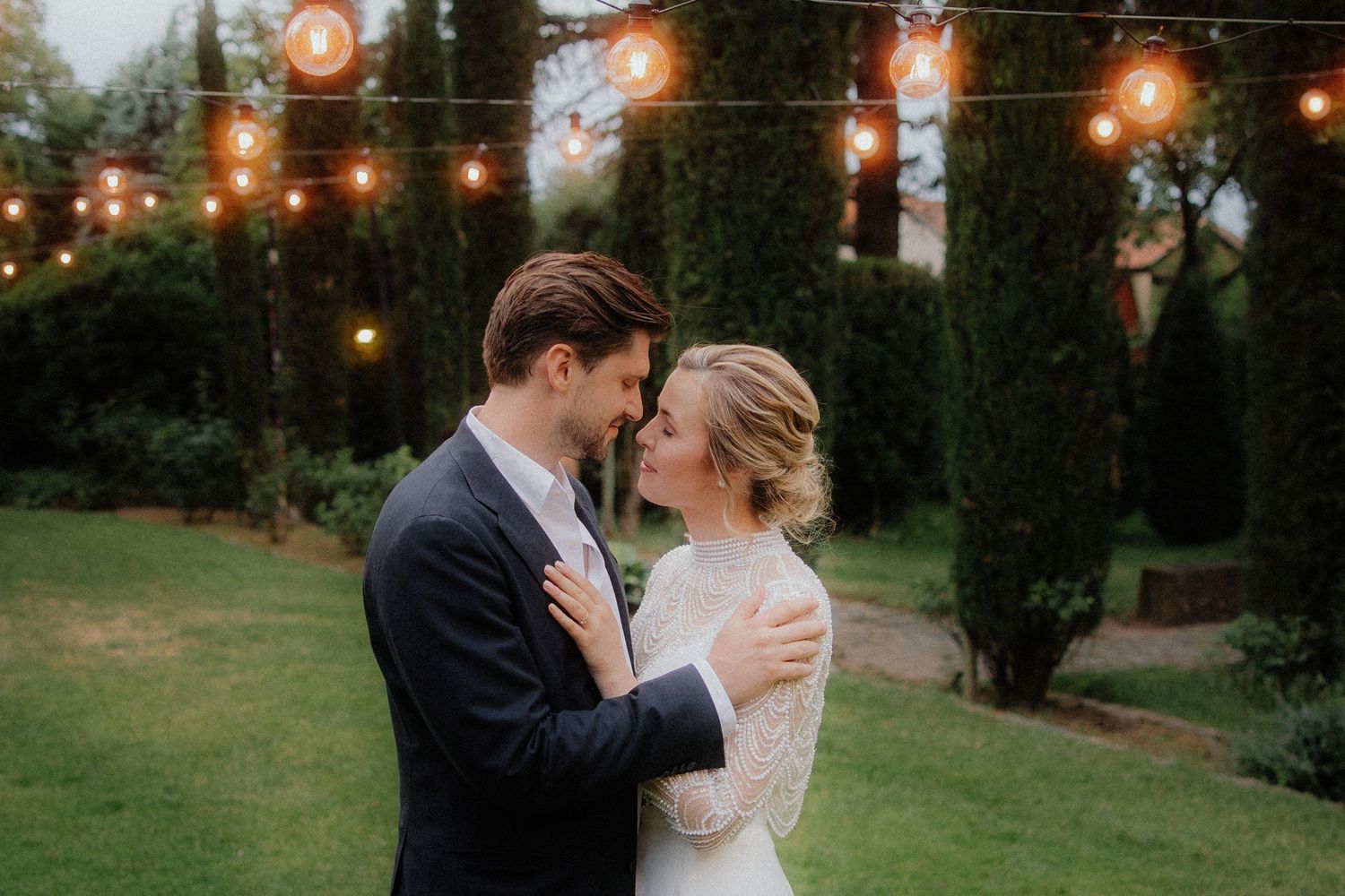 A bride and groom share a romantic moment outdoors under warm string lights surrounded by tall cypress trees at dusk.
