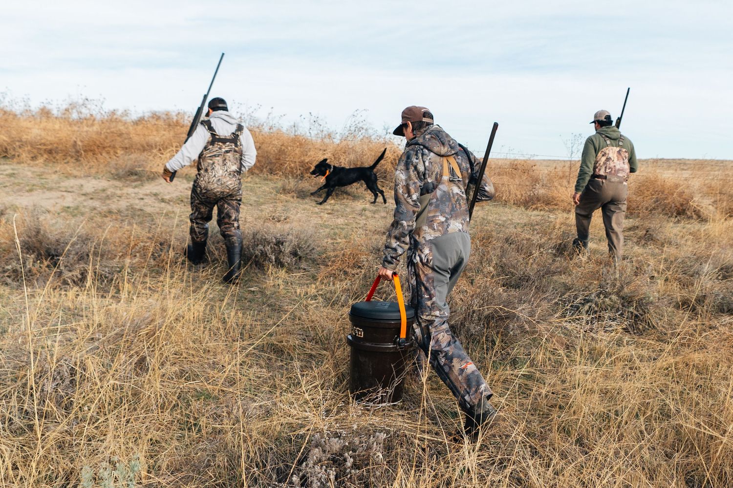 Three hunters in camouflage walk through dry grassland with a black dog during a hunting expedition.