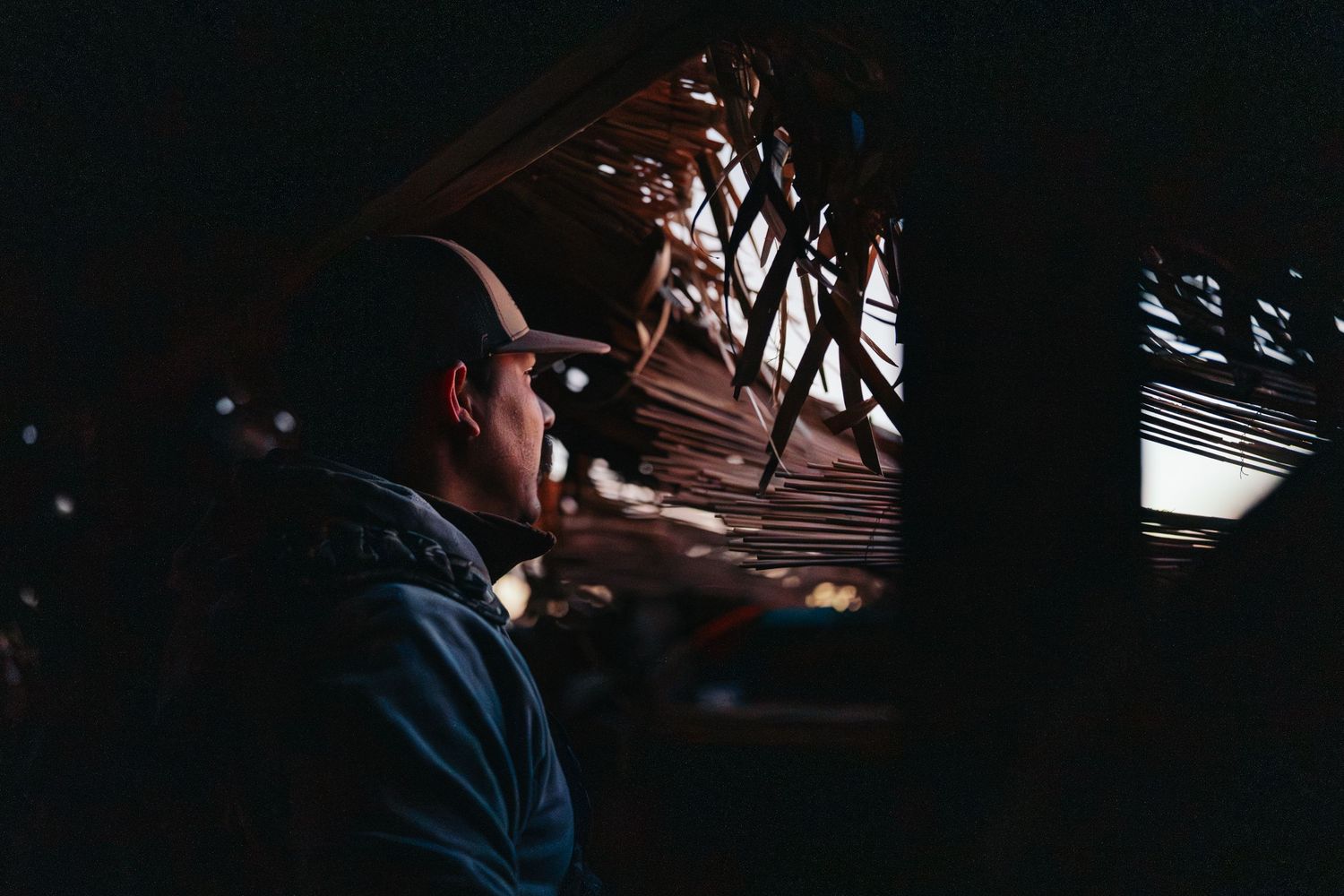 Man in hat gazes upward in dark rustic setting with woven bamboo ceiling structure overhead.