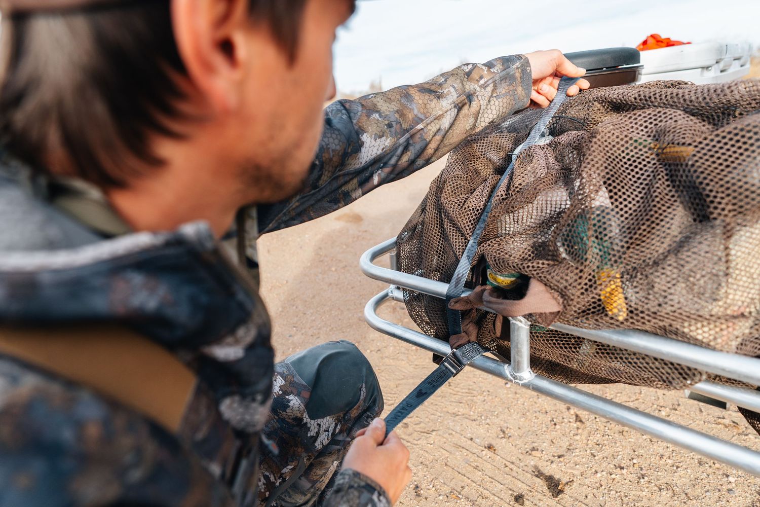 Hunter in camouflage gear handling a fishing net with caught fish near a metal frame outdoors.
