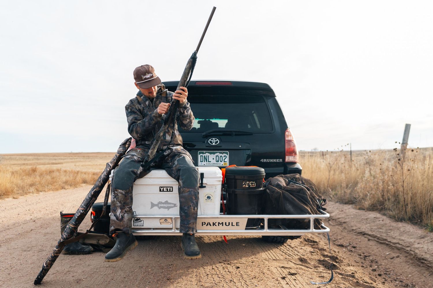 Hunter sitting on truck tailgate holding rifle in rural field, wearing camo gear and hat near Toyota SUV.