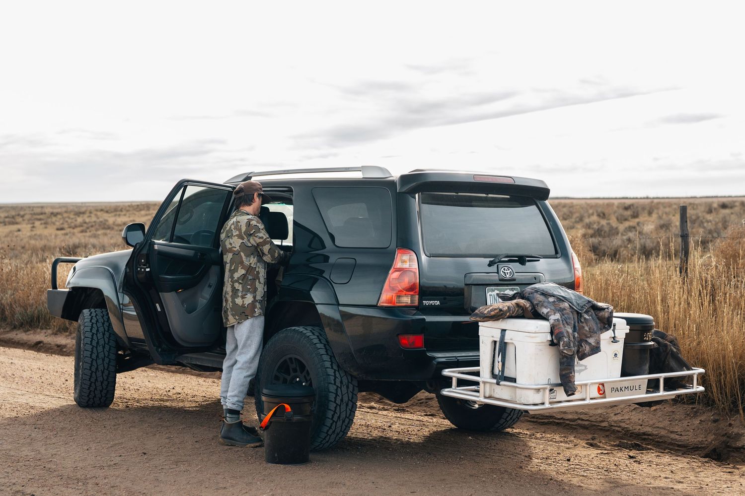 Man loading gear into a black Toyota 4Runner SUV with a rear cargo carrier on a dirt road in open grasslands.