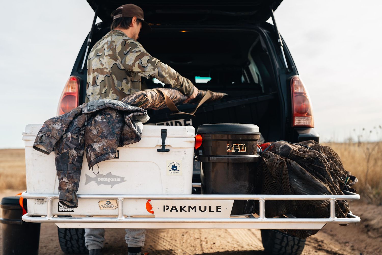 Hunter in camo gear loading gear into a truck bed with a Pakmule rack and cooler in an open field.