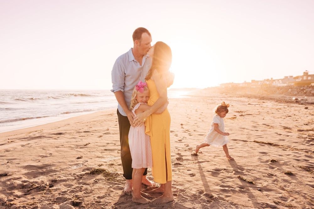 Family embracing on a sunny beach at golden hour with children playing in the sand by the ocean.