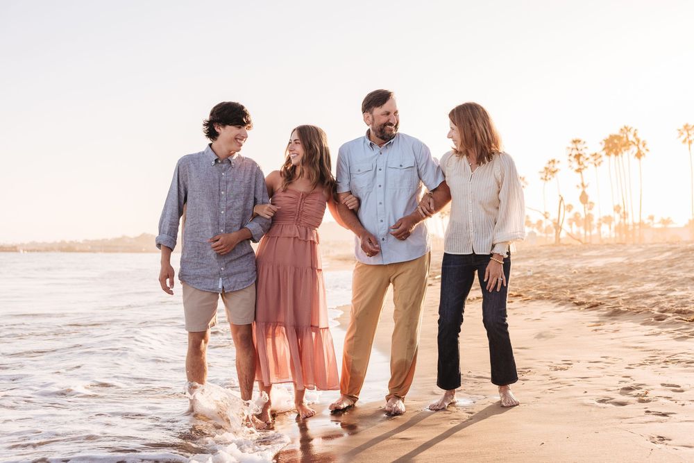 Family of four laughing and walking along a sunny beach shoreline at golden hour, waves splashing at their feet.