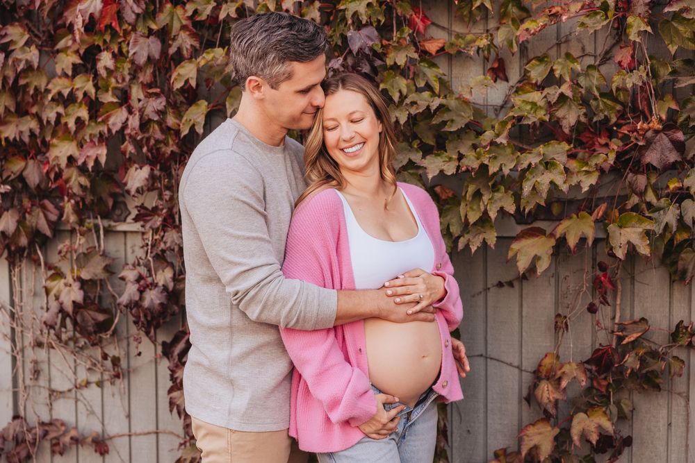 Smiling pregnant couple embracing outdoors in autumn, woman in pink cardigan cradling baby bump near red ivy-covered fence.