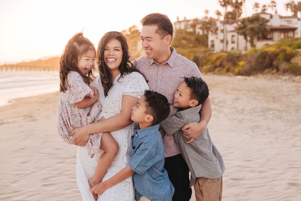 Happy Asian family of five sharing a warm embrace on a sunny beach at golden hour.