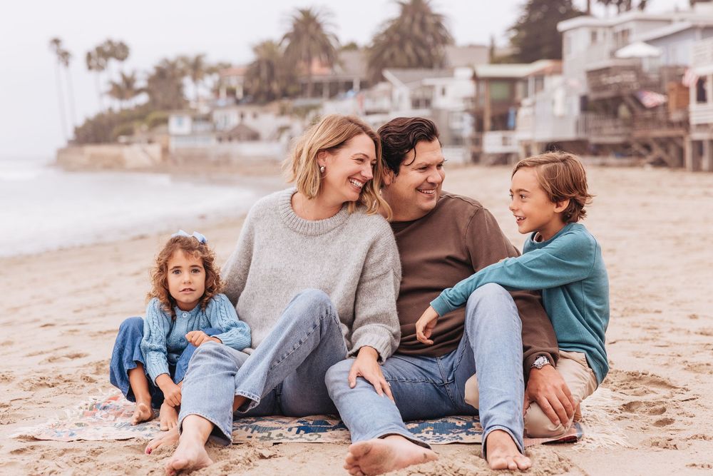 Happy family of four sitting together on a sandy beach, smiling and enjoying quality time near the ocean.