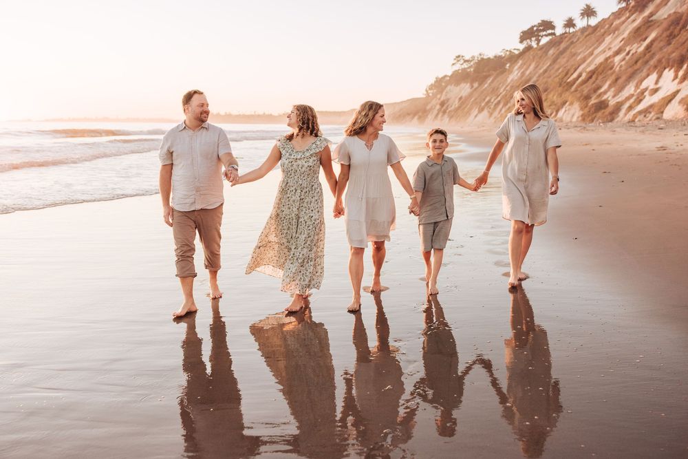 Family of five holding hands walking on a reflective wet beach at sunset near cliffs.