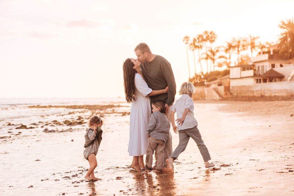 Family of five sharing a tender moment at sunset on a tropical beach, parents kissing while children play in the sand.
