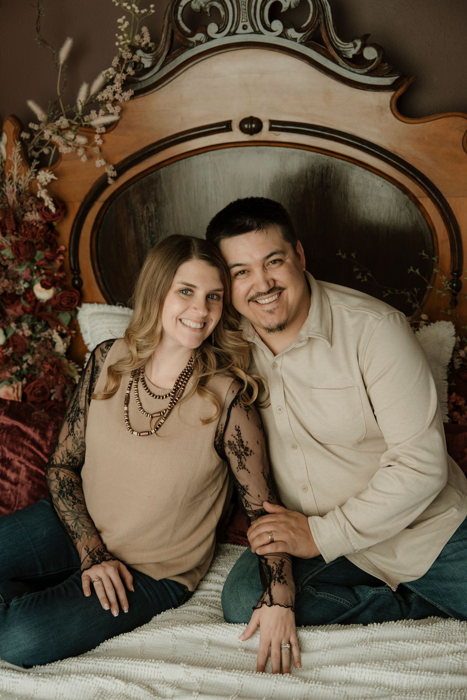 Smiling couple sitting together on a bed with ornate headboard and floral decor in a cozy studio portrait.