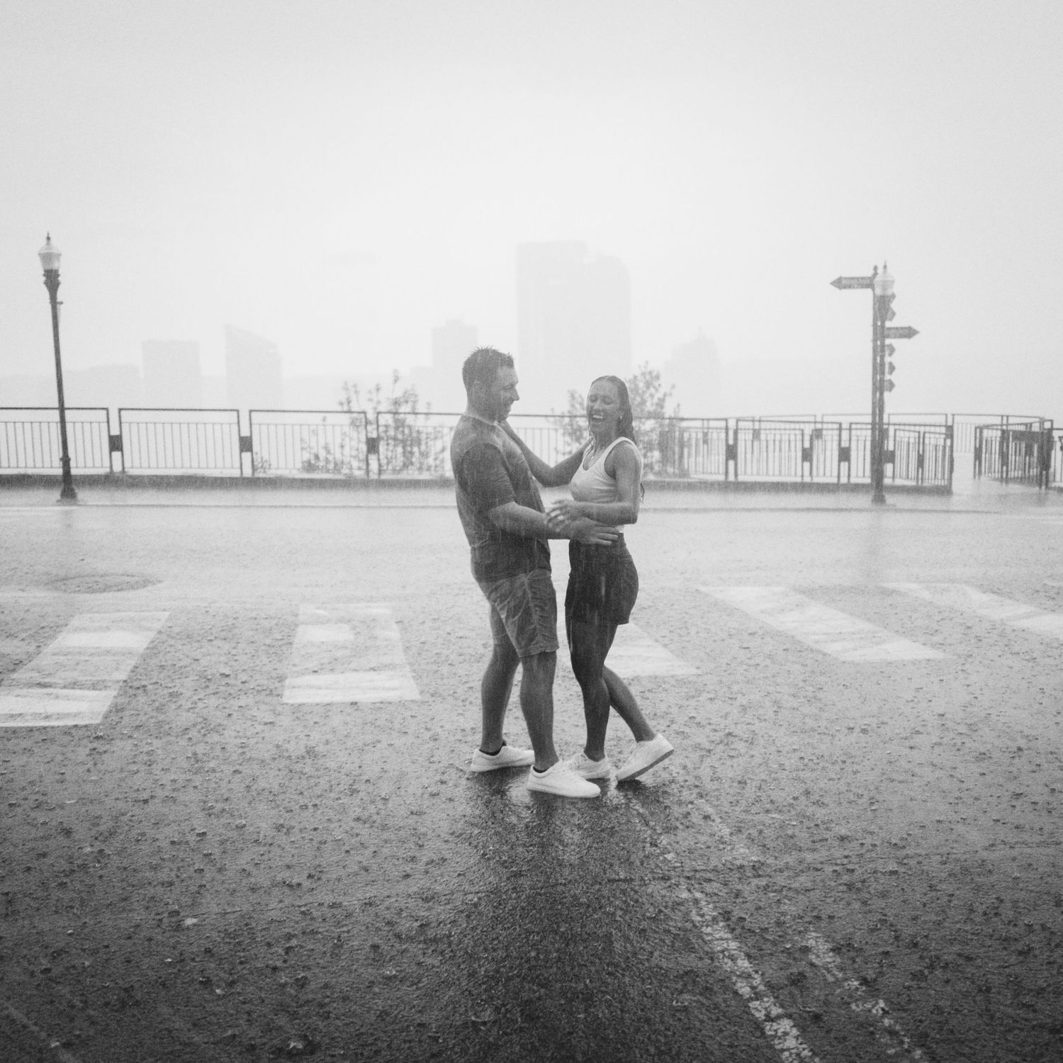 Black and white photo of a couple dancing together in the rain on a wet city street with foggy skyline.