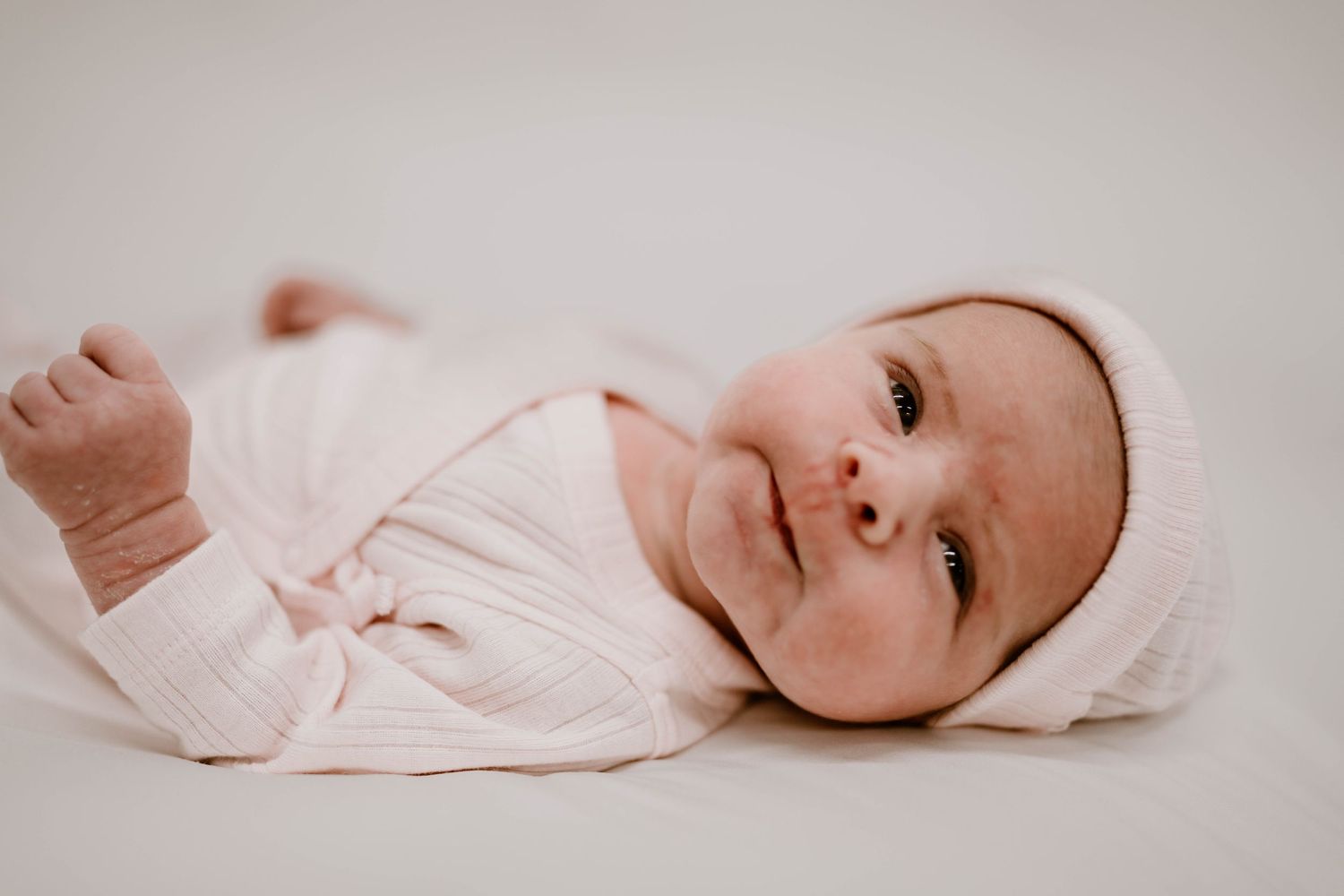 Newborn baby in pink outfit and hat lying on white blanket, looking at camera with bright eyes.