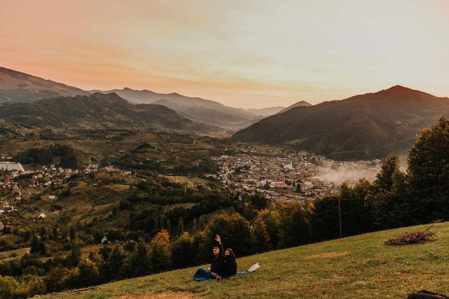 Couple sits on hillside overlooking misty mountain valley town at golden hour sunset.