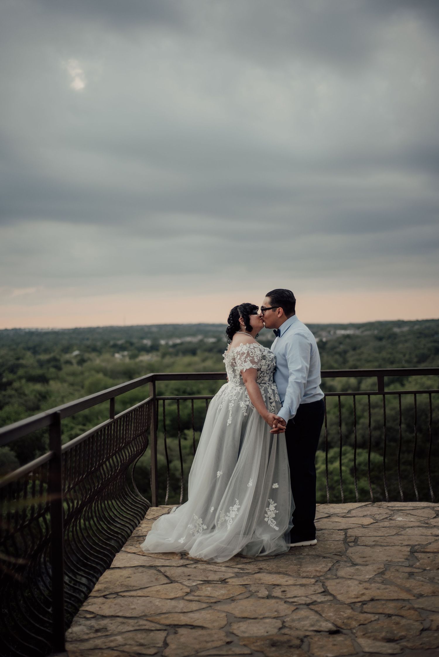 A romantic wedding couple embraces on a stone overlook with sweeping countryside views under moody evening skies.