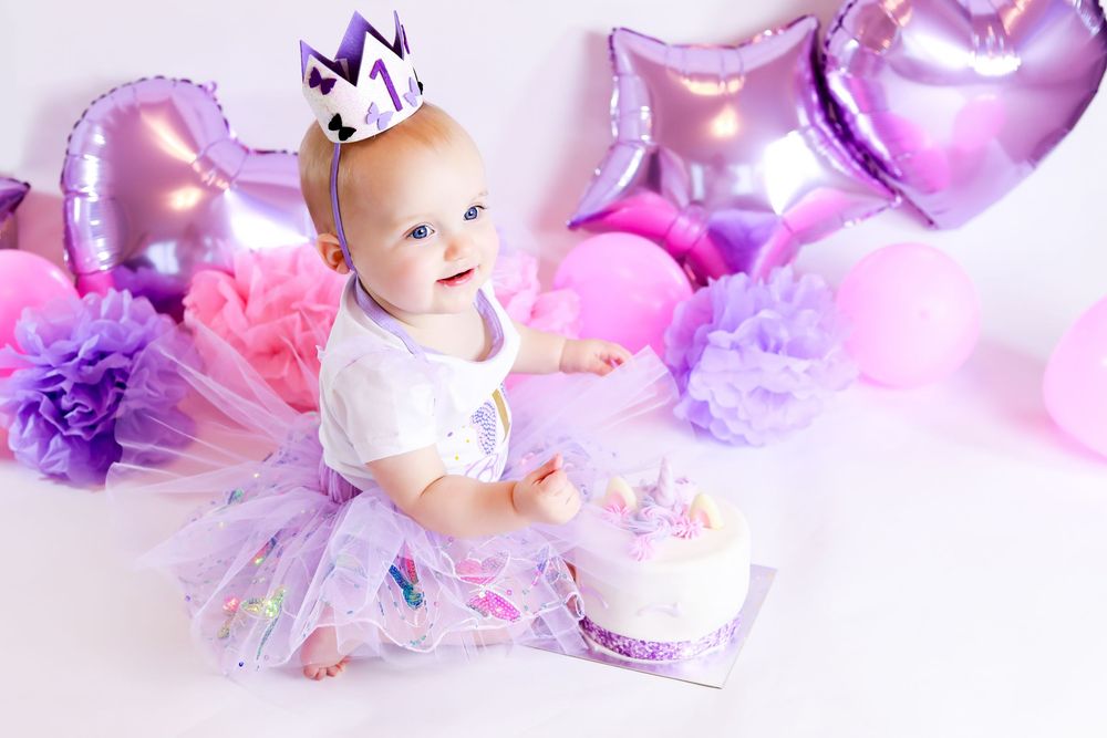 Baby in purple tulle dress with crown sitting next to cake and metallic heart balloons.