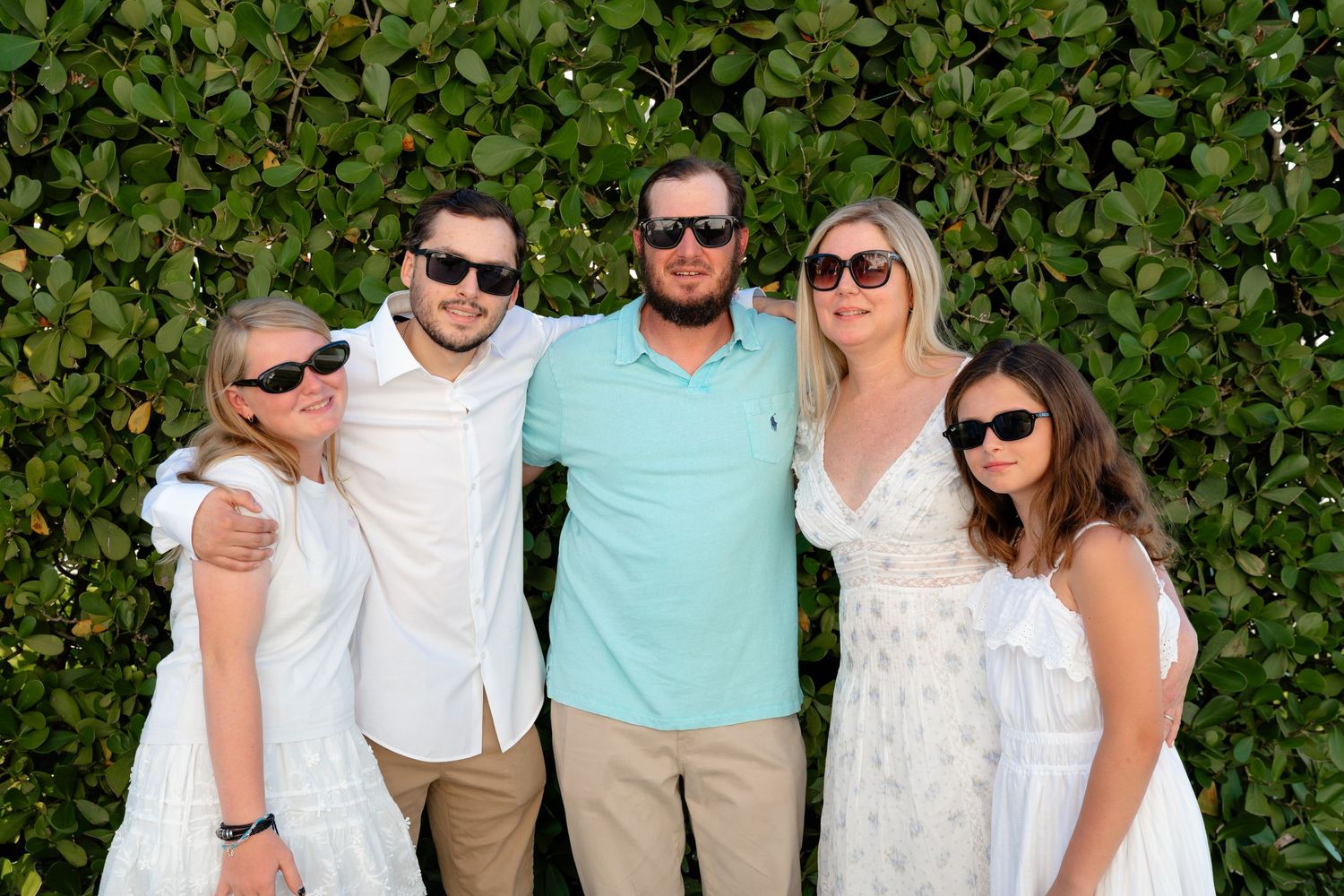 A couple holding hands in front of a green hedge, man in blue shirt and khakis, woman in white sundress.