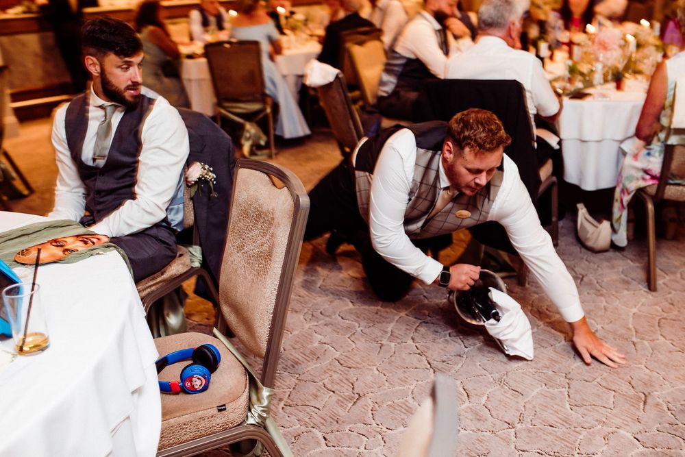 Two groomsmen in vests at a wedding reception, one kneeling on the floor picking up items near an open suitcase.