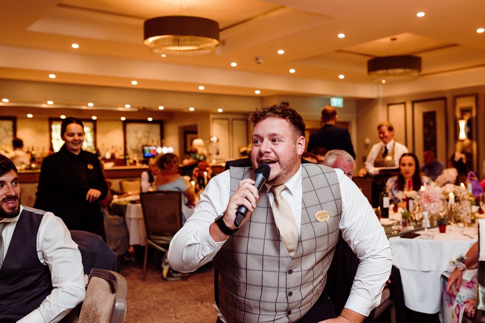 Man in plaid vest singing into microphone at wedding reception with guests seated at decorated tables.