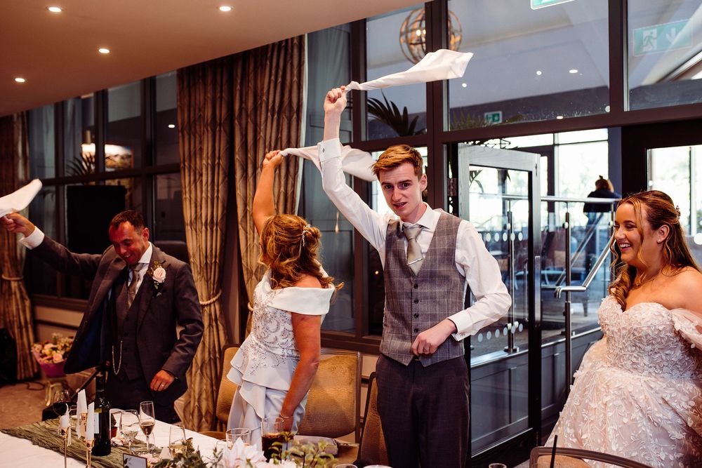 Groom cuts wedding cake while bride and guests wave napkins at a festive indoor wedding reception.