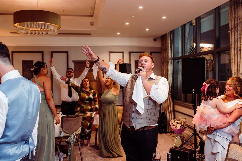 A male singer performs energetically at a wedding reception, holding a microphone while guests dance and celebrate around him.