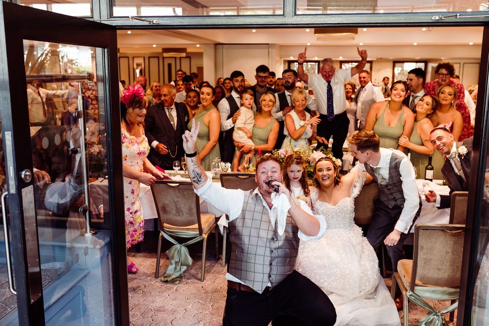 Joyful bride and groom celebrate at wedding reception with smiling guests gathered behind them in elegant venue.