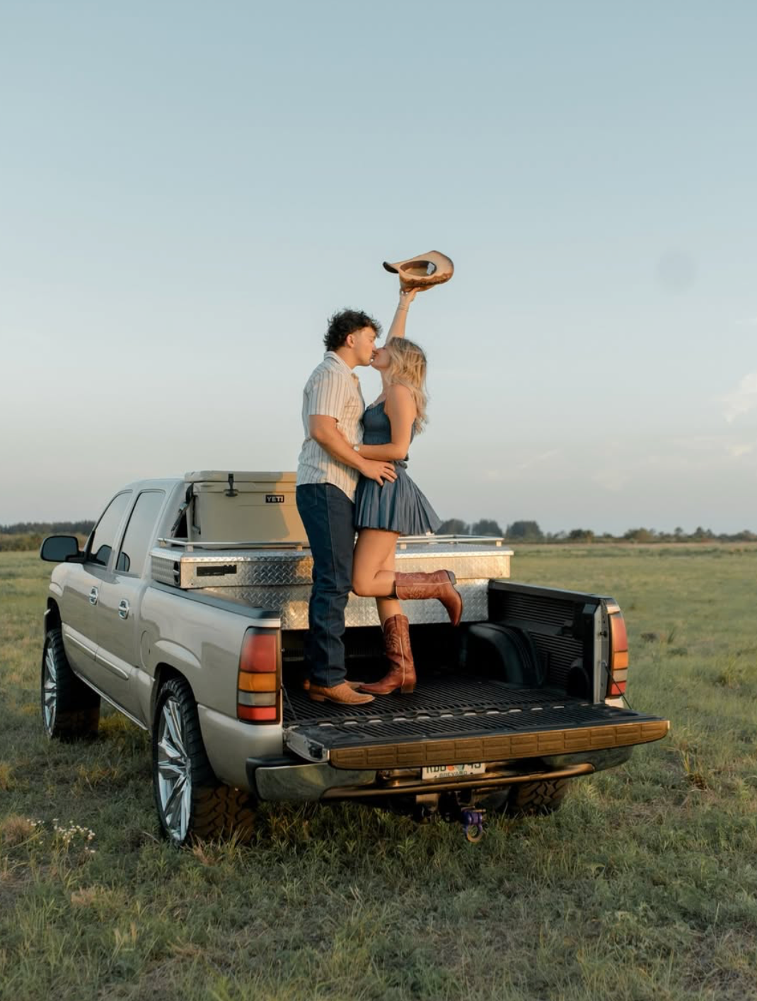 Couple kissing in truck bed holding cowboy hat in open field at golden hour, country engagement photo.