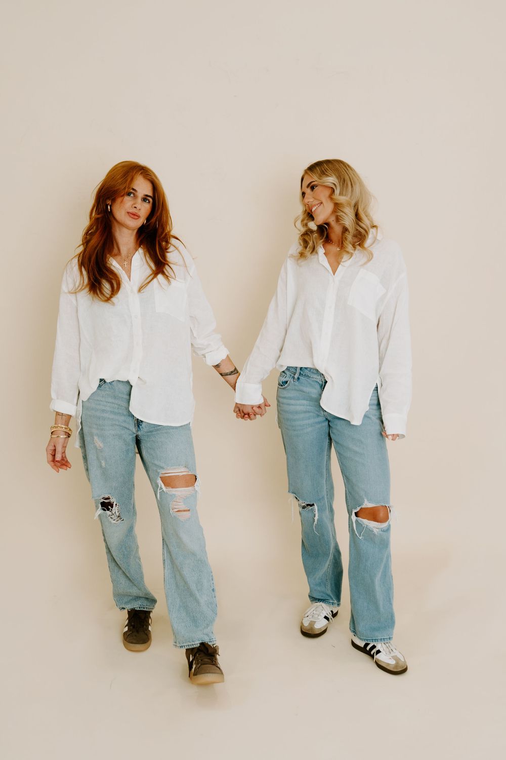 Two women in matching white button-up shirts and ripped light-wash jeans hold hands against a beige background.