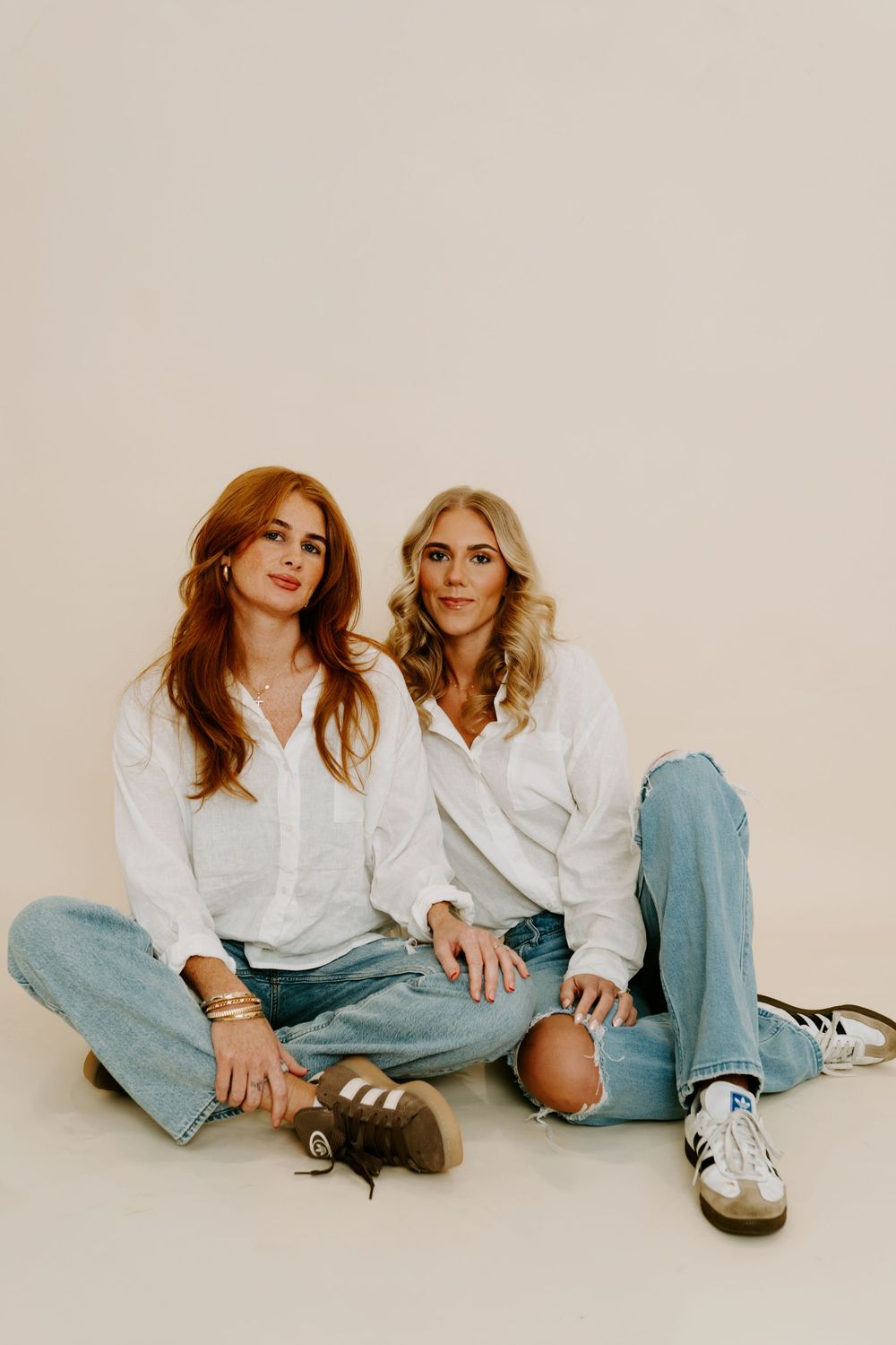 Two women in white shirts and jeans sitting together on a beige background, smiling at the camera.