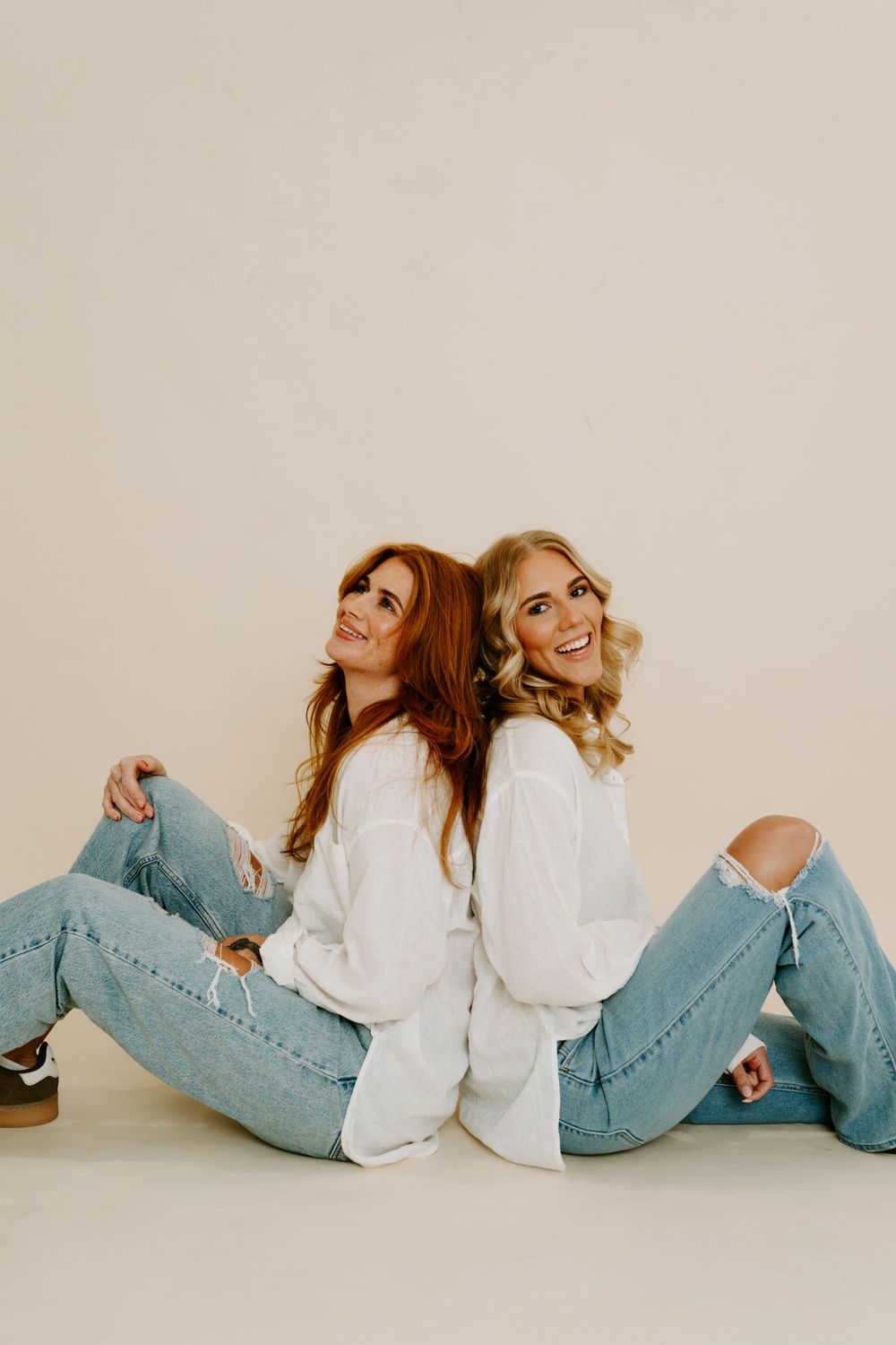 Two smiling women in white tops and ripped jeans sit back-to-back on a light beige studio background.