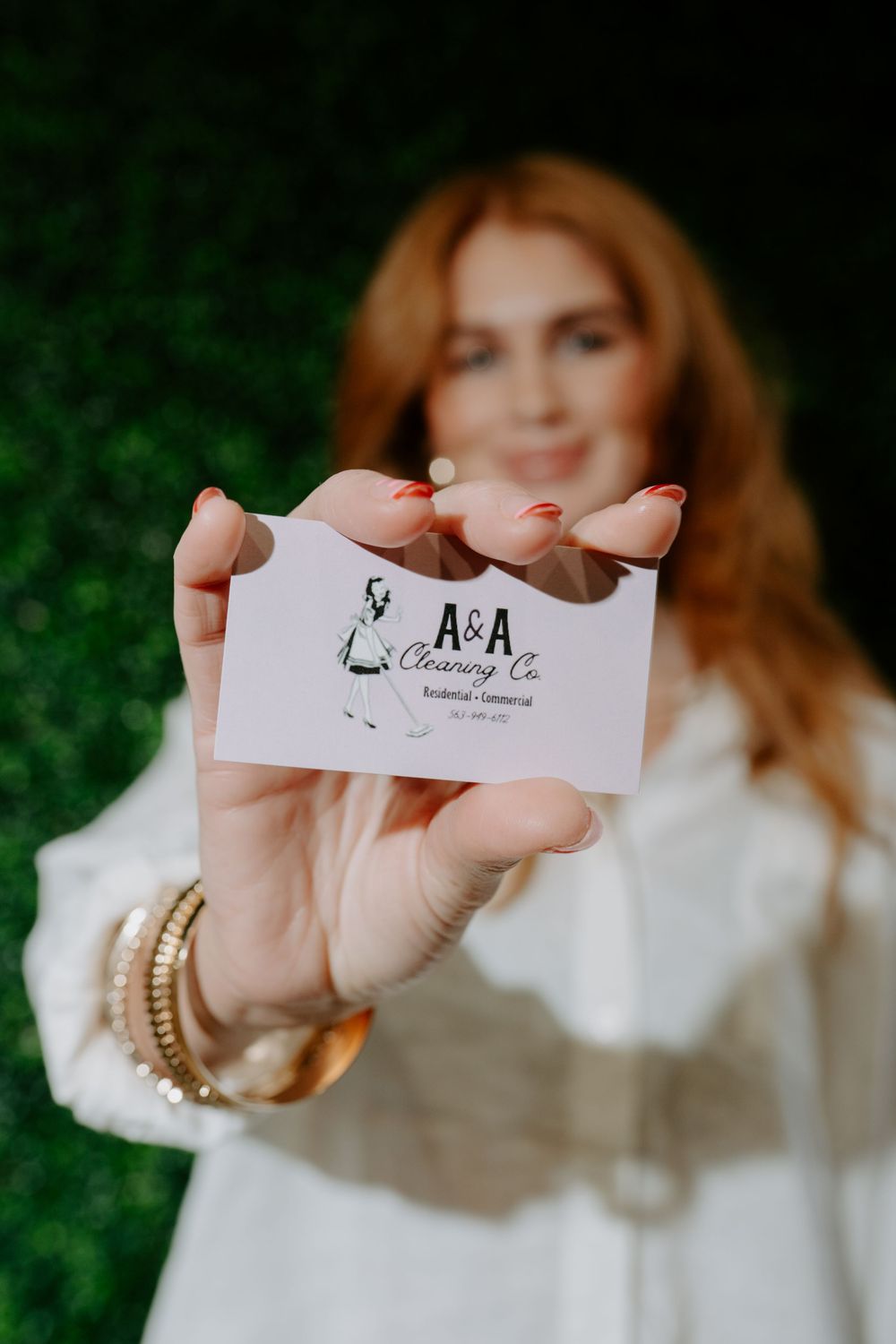 Red-haired woman in white blouse holding A&A Cleaning Co. business card toward camera outdoors.