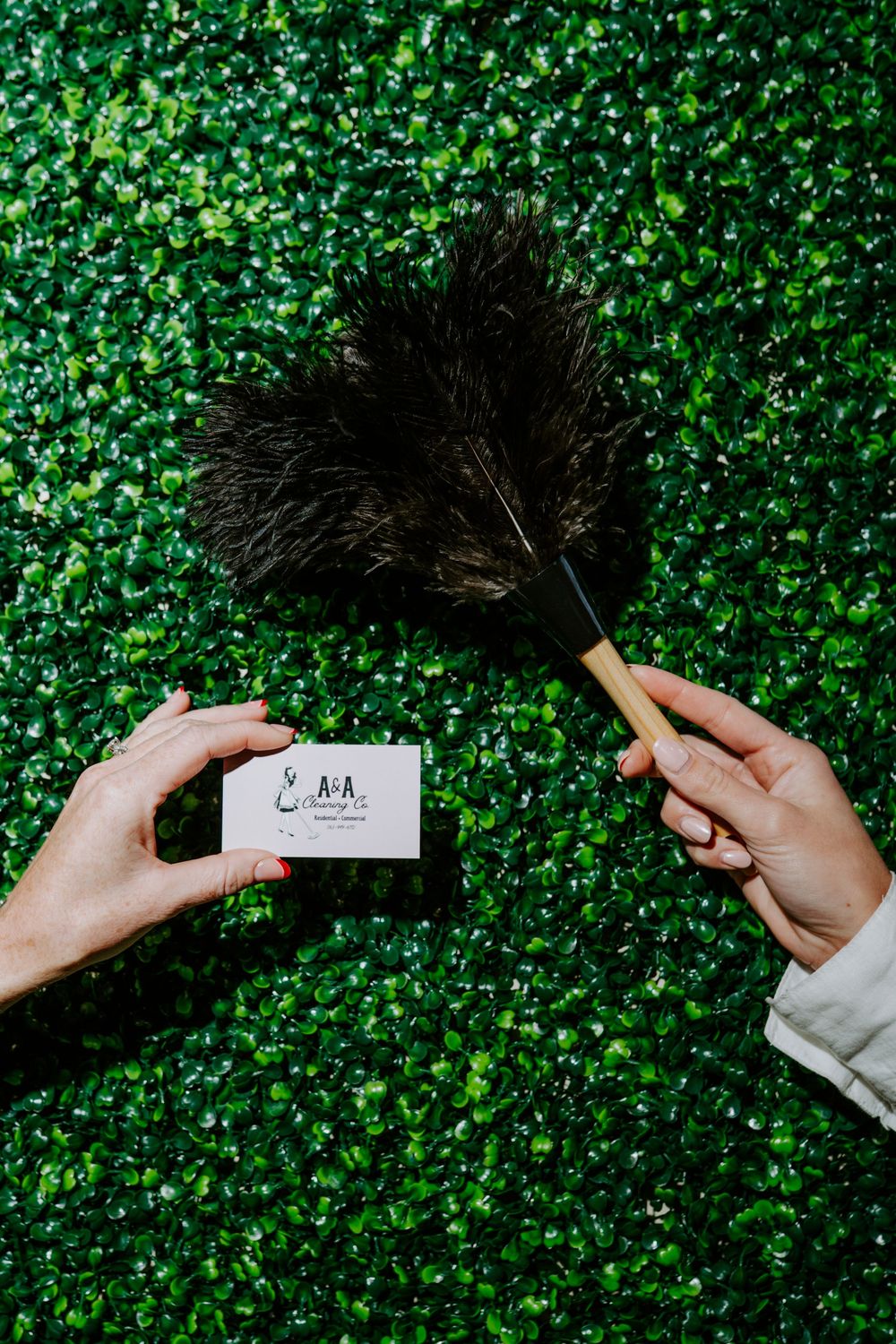 Hands holding a business card and feather duster against a green hedge wall for A&A Cleaning Co.