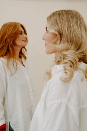 Two women in white linen shirts, one blonde and one redhead, facing each other in a bright, minimalist setting.