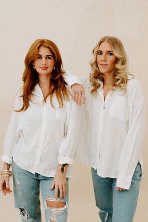 Two women in white button-up shirts and ripped jeans pose together against a beige background.