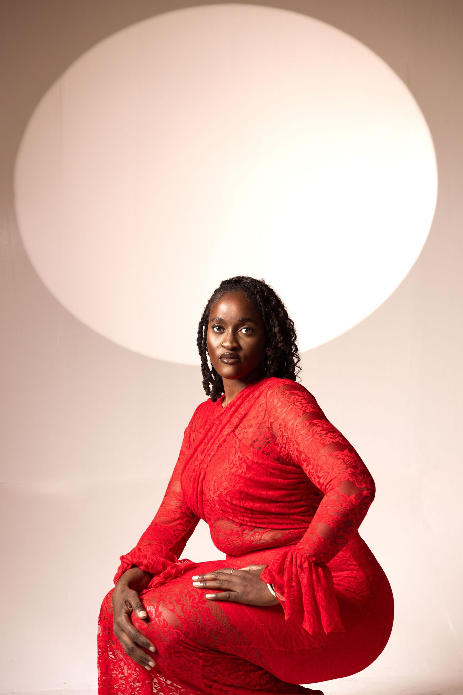 A woman in a red lace dress sits gracefully in front of a circular white light backdrop.