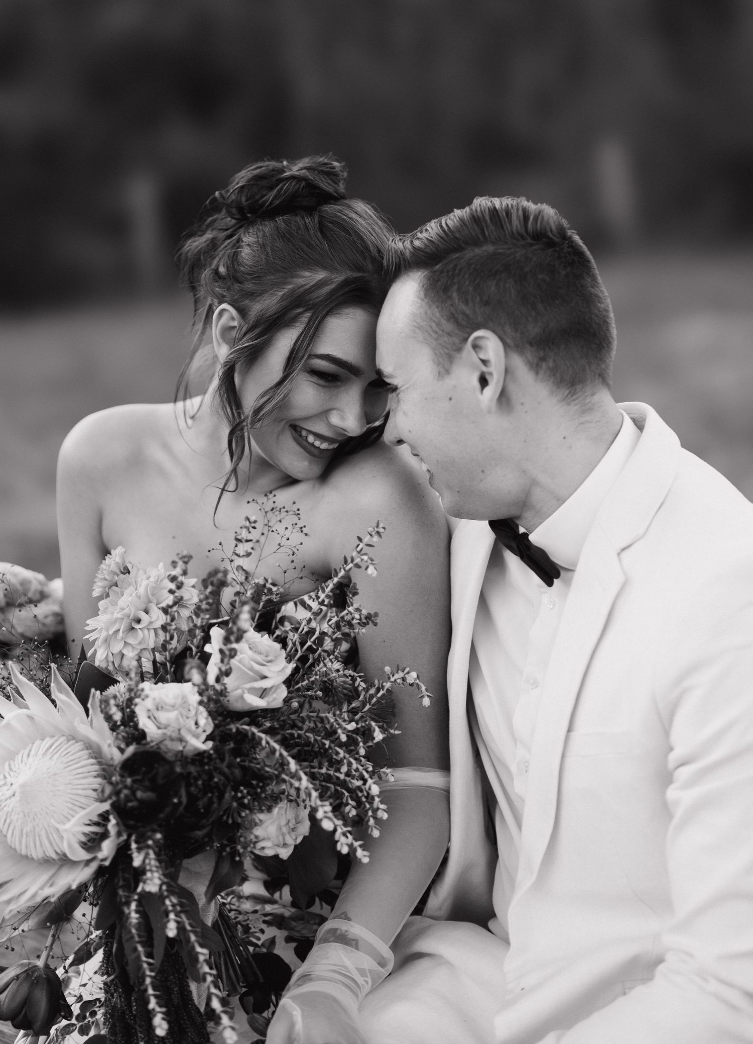Black and white photo of a smiling bride holding a bouquet leaning close to a groom in a white suit.